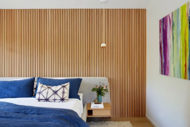 A contemporary bedroom with a large vertical timber slat feature wall behind the bed, a low upholstered headboard, layered blue and white bedding, and a compact timber bedside table. A colourful abstract artwork adds a vivid accent on the white wall, while a small pendant light and neutral rug complete the calm, refined interior.