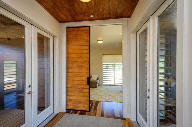 A bright residential entry hall with white-framed glazed doors on both sides, a large timber pivot door, warm timber ceiling lining, and polished timber flooring. The hall opens toward a carpeted living or bedroom area with horizontal window screening. The space has a clean, contemporary coastal feel with strong natural timber detailing.