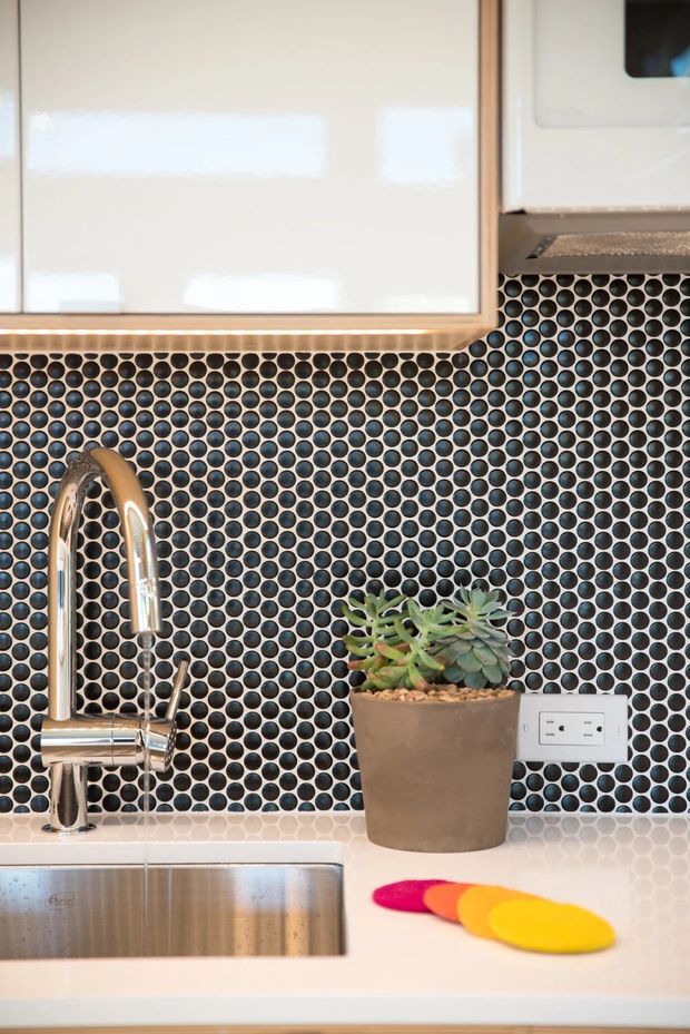 Close-up view of a compact kitchen work area featuring a stainless-steel sink and curved chrome faucet, a white engineered-stone countertop, glossy white cabinetry with timber edging, and a striking backsplash of small circular dark blue-grey mosaic tiles. A potted succulent and brightly coloured round coasters sit on the counter.