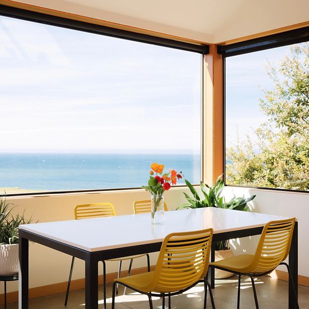 A bright dining area with a rectangular white dining table and mustard-yellow slatted chairs, positioned beside expansive corner windows overlooking the ocean. Warm timber window frames, indoor plants, and a vase of flowers add natural detail to the airy coastal setting.