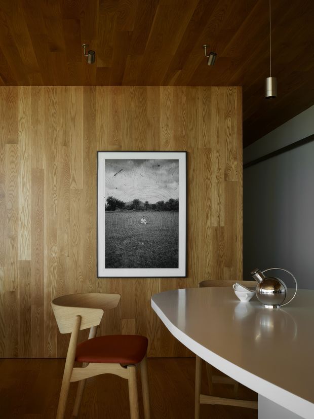 Warm, moody interior detail featuring a wood-lined ceiling and full-height timber feature wall. A framed black-and-white photograph hangs above a sculptural wooden chair with a rust-red upholstered seat, while the foreground is dominated by a rounded white dining or work table with a metallic desk lamp. Dark grey walls and carefully integrated track and pendant lighting create a refined contemporary atmosphere.