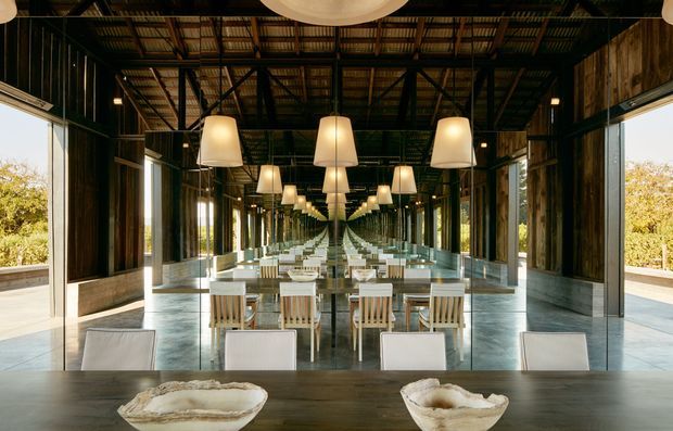 A dramatic, symmetrical dining space with a long dark timber dining table, pale upholstered chairs, sculptural pendant lights, full-height mirrored walls, and extensive glazing opening to a leafy landscape. The exposed dark structural frame and corrugated metal ceiling create an industrial warehouse-like character, while the polished concrete floor and repeated reflections emphasize the room's depth.