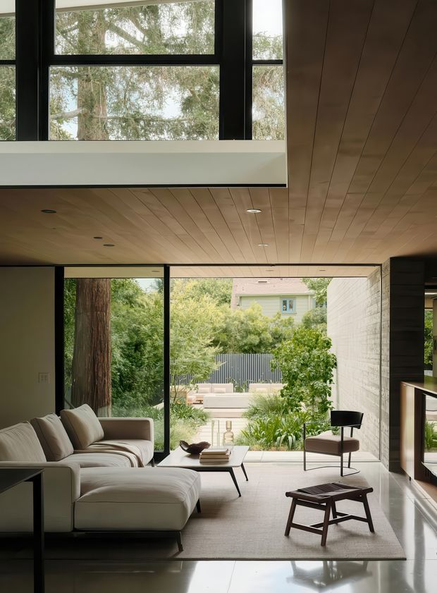 A refined contemporary living room with expansive black-framed glazing opening onto a lush garden courtyard. The room features a large cream sectional sofa, minimalist lounge chairs and coffee tables, a polished light floor, a warm timber-lined ceiling, and a white masonry feature wall. A high-level window brings additional daylight and creates a strong indoor-outdoor connection.