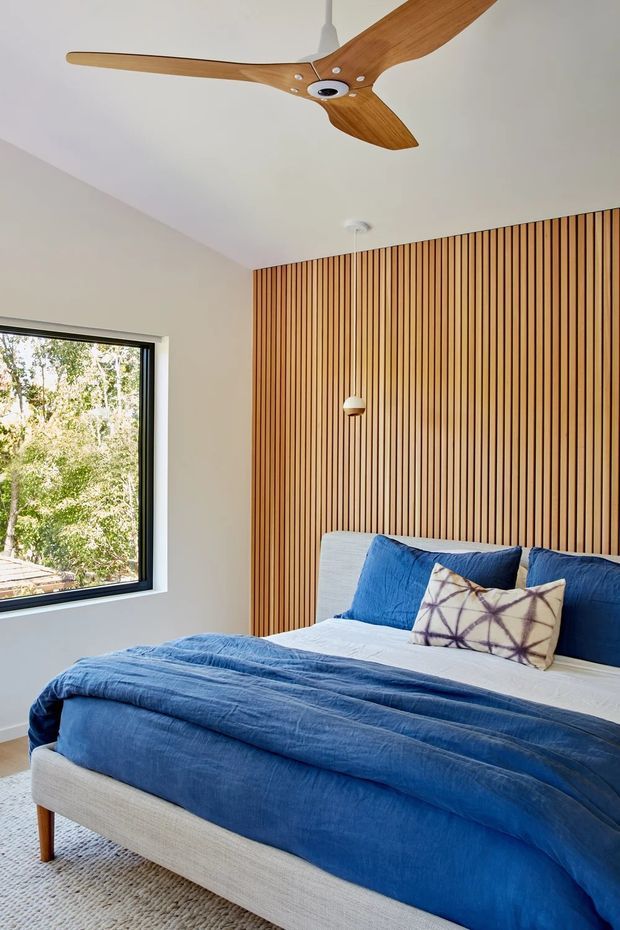 A bright, contemporary bedroom with a large upholstered bed dressed in layered blue bedding and cushions. The feature wall is clad in vertical warm timber battens, complemented by a white ceiling, black-framed window, minimalist pendant light, and a timber three-blade ceiling fan. Trees are visible outside the window.