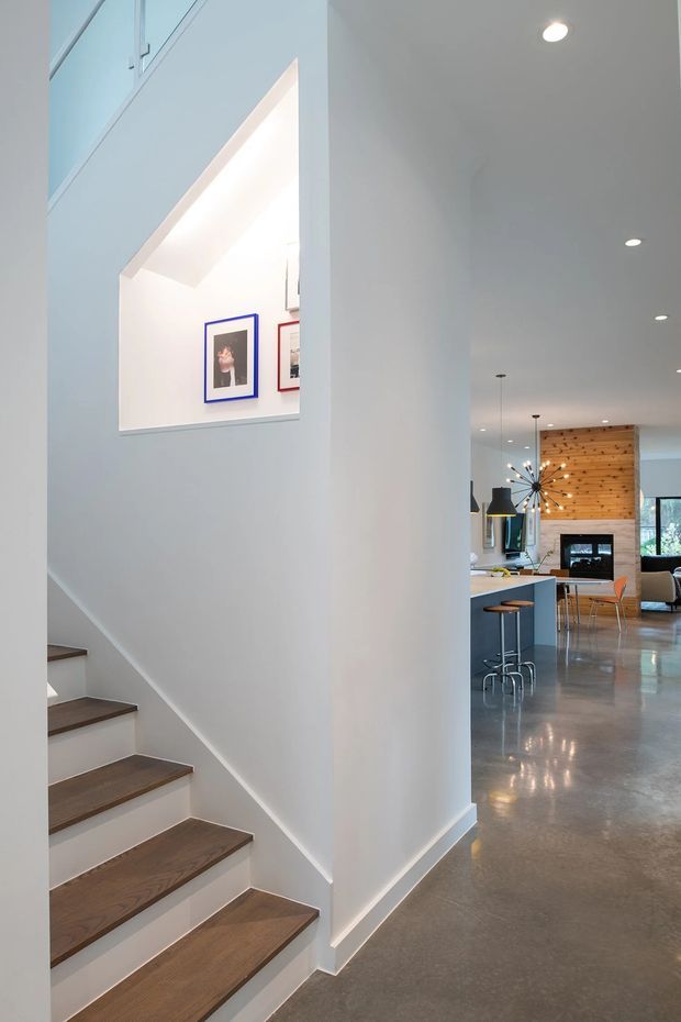 Bright contemporary open-plan home interior viewed from a stairway, with white walls, a timber-tread staircase, polished concrete flooring, recessed ceiling lights, and a glimpse of a kitchen and living area beyond. The open stair void and framed artwork add architectural interest, while a timber feature wall and modern fireplace are visible in the background.