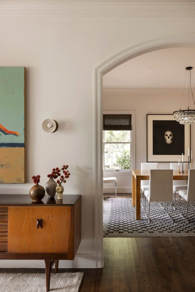 A refined interior view from a hallway or entry area into a formal dining room through a wide arched opening. The foreground features a mid-century timber credenza with sculptural ceramic vases and artwork, while the adjoining dining room has a wooden table, upholstered dining chairs, patterned rug, framed artwork, chandelier, and a window with a woven blind. Walls and ornate architraves are painted in warm off-white tones, with dark timber flooring throughout.