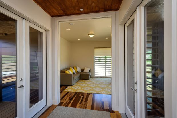 A bright residential interior looking through a glazed passage or entry area into a compact living room. The space features extensive white-framed glazed doors, warm timber flooring, a timber-lined ceiling, cream walls, a patterned area rug, and a small sectional sofa with yellow and neutral cushions. Horizontal screened glazing at the far end provides filtered daylight.