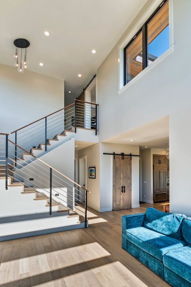 Bright contemporary open-plan entry and living area featuring a prominent staircase with slim dark metal balustrades and horizontal cables, warm timber handrails and stair treads, pale walls, timber flooring, tall windows, and a vivid blue sectional sofa. A high ceiling and open stair void create an airy, spacious feel, with a kitchen and timber double doors visible beyond.