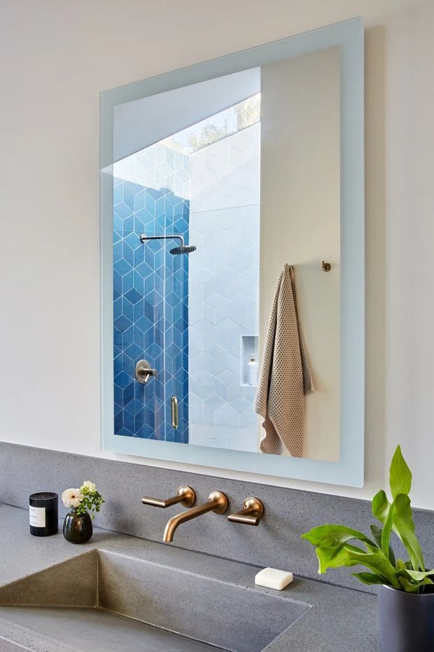 A contemporary bathroom vanity viewed straight on, featuring a large pale blue-framed mirror, a grey concrete-look integrated basin and backsplash, and brushed brass wall-mounted tapware. The mirror reflects a walk-in shower lined with blue and white geometric tiles, with a beige textured towel hanging beside it. Small toiletries and a leafy potted plant sit on the vanity.