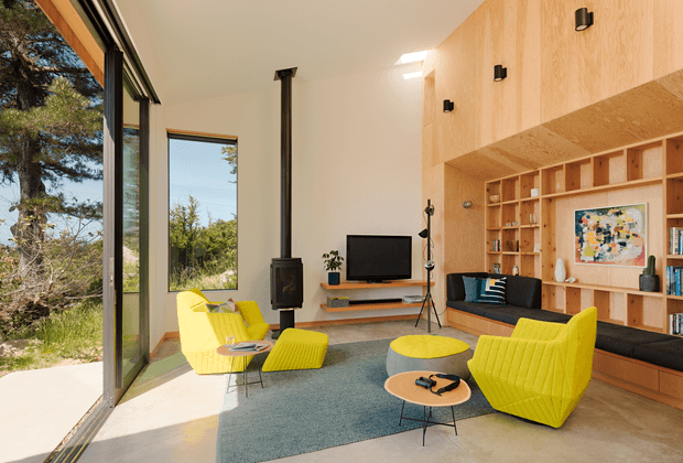 Bright contemporary living room with floor-to-ceiling glazing opening toward a landscaped setting. The room features a built-in timber wall and shelving unit, pale timber ceiling and wall surfaces, a compact black wood-burning stove, television, dark built-in window seat, blue area rug, and vivid yellow lounge chairs with a matching ottoman. A low round coffee table and large artwork complete the warm, playful interior.