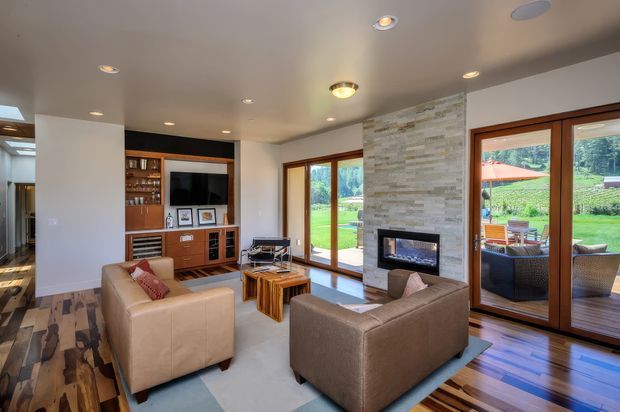 Contemporary living room with a warm neutral palette, timber flooring, large timber-framed glazed doors and windows, a stone-clad fireplace wall, built-in timber entertainment cabinetry, and seating arranged around the fireplace. The glazing opens onto a landscaped outdoor terrace and garden.