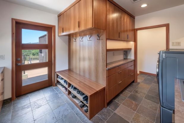 A compact, well-organised mudroom or entry area with extensive warm timber built-in cabinetry. The space includes a timber bench with open shoe storage below, coat hooks on a vertical timber panel, overhead cupboards, and a long run of drawers and shelving. A glazed timber-framed exterior door opens to a deck, while dark slate-look floor tiles and a large blue appliance or cabinet are visible at right.