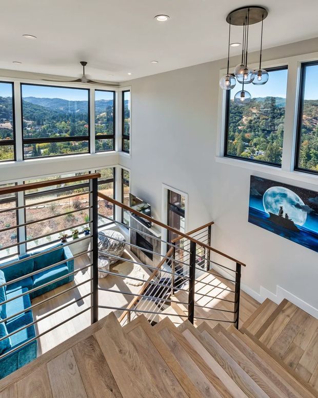 A bright, contemporary multi-level stairway with broad engineered-wood treads, slender dark metal balustrades, and timber handrails. Floor-to-ceiling corner glazing overlooks a wooded hilly landscape, while a sculptural cluster pendant hangs above the stair void. A lower living area with blue seating is visible below, along with wall art and recessed lighting.