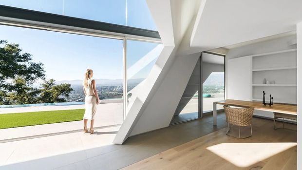 A bright, contemporary open-plan dining and living area with floor-to-ceiling glazing opening to a terrace and pool. The room features white built-in shelving, a long timber dining table with woven chairs, pale timber flooring, strong geometric structural elements, and expansive views beyond the property.