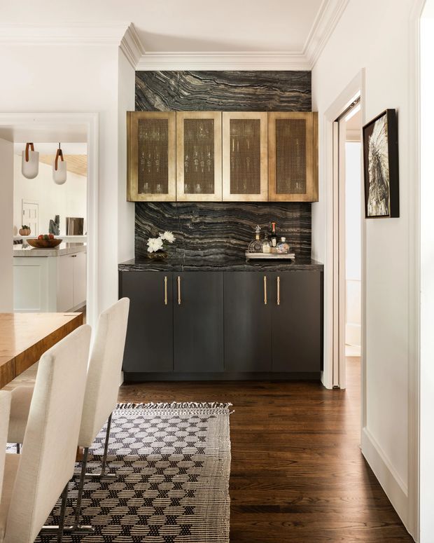 Elegant transitional interior vignette featuring a dark built-in sideboard with brass hardware, textured woven-front upper cabinets, and a dramatic dark veined stone feature wall. Warm timber flooring, a patterned monochrome rug, classic white wall mouldings, and a partially visible dining area create a refined, layered hallway or dining space.