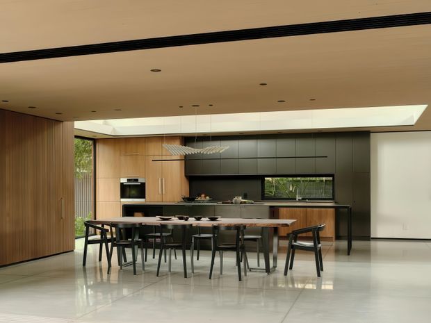A refined open-plan kitchen and dining area with a long timber dining table and black-framed chairs in the foreground. The kitchen features dark matte cabinetry, timber wall panelling and cabinetry, integrated appliances, a dark benchtop, and a horizontal window. A large linear sculptural pendant hangs above the table, while a broad ceiling skylight and full-height glazing bring in natural light. The space has a restrained contemporary architectural interior with polished light flooring and strong black detailing.