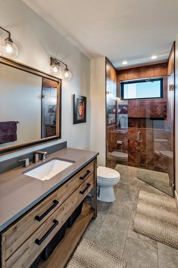 A well-appointed bathroom with a long timber vanity, grey stone-look benchtop, undermount sink, large framed mirror, toilet, and a walk-in glass shower enclosure. The shower features warm brown tiled walls with a horizontal decorative band, a high window, and recessed ceiling lights. Neutral grey floor tiles, black hardware, and exposed-bulb wall lighting complete the space.