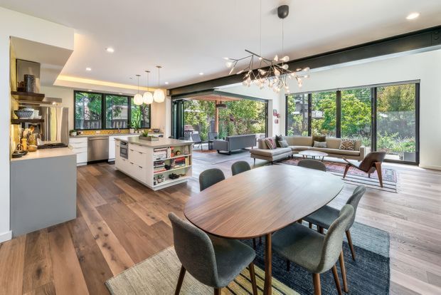 Open-plan contemporary living, dining and kitchen interior with extensive floor-to-ceiling glazing opening toward a leafy outdoor setting. The space features warm timber flooring, a white kitchen with central island and integrated appliances, a large oval timber dining table, upholstered dining chairs, a lounge area with sofas and occasional chairs, recessed ceiling lighting, pendant lights and a sculptural chandelier. Black-framed glazing and doors create a strong connection to the garden.