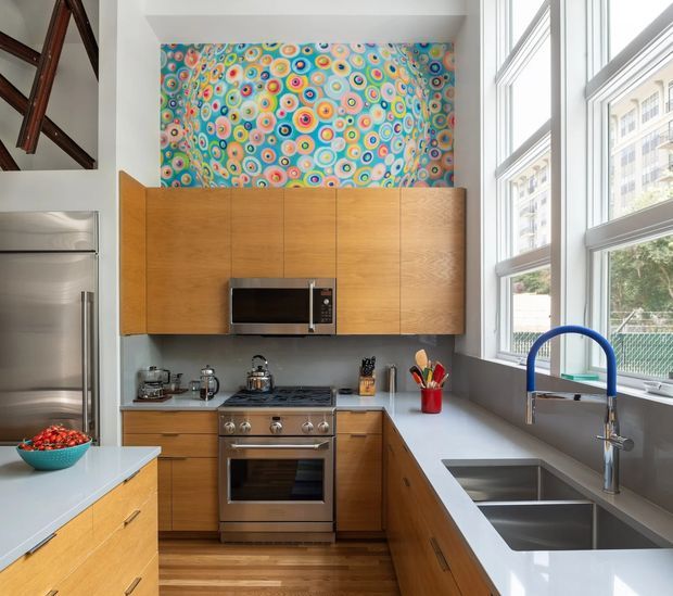 A bright, compact contemporary kitchen with warm wood cabinetry, white benchtops, stainless-steel appliances, a large double-bowl undermount sink, and a distinctive blue gooseneck tap. Floor-to-ceiling windows bring in abundant daylight and overlook an urban setting. A colourful patterned feature artwork or backsplash panel adds a playful accent above the upper cabinets.