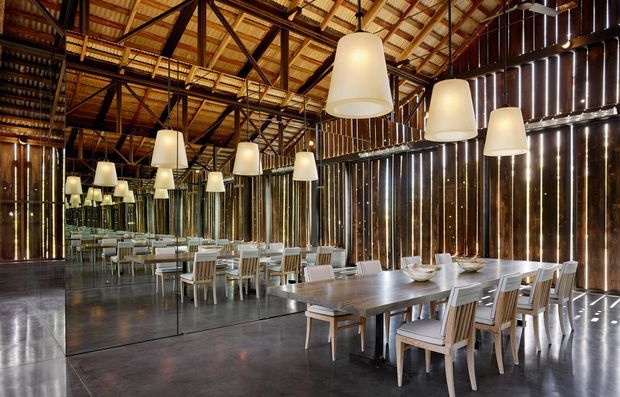 A spacious dining area with long timber dining tables and upholstered chairs, illuminated by multiple oversized pendant lights. Full-height mirrored panels reflect the room, while dark vertical timber wall cladding and exposed timber-and-steel roof trusses create a dramatic industrial interior. The polished dark floor enhances the warm, refined atmosphere.