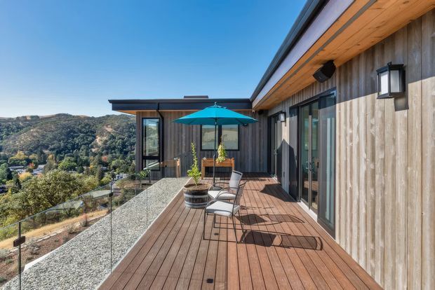 A sunny elevated timber-clad house with a spacious outdoor terrace overlooking a wooded hillside valley. The terrace features timber decking, frameless glass balustrades, a turquoise umbrella, outdoor chairs, and potted plants. The building has a low-pitched roof, extensive glazing, vertical weathered timber cladding, and warm timber soffits.