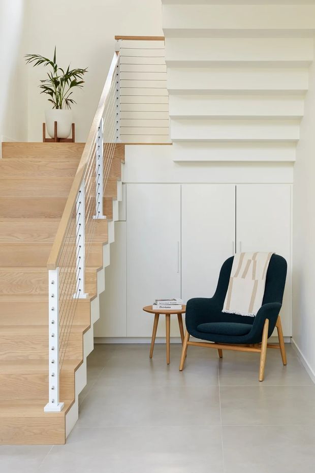 Bright minimalist stairway and landing with pale timber open-riser stairs, white painted balustrade and cable railing, built-in white cabinetry beneath the stairs, and a dark teal upholstered lounge chair with a small timber side table. A potted plant sits on the upper landing, and the space has a clean, contemporary feel with large pale grey floor tiles.