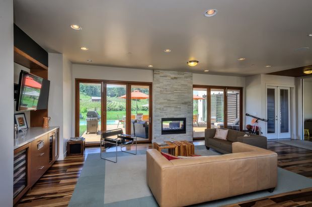 A contemporary living room with a large tan sectional sofa, glass coffee table, built-in dark timber media cabinetry, recessed lighting, and a central stacked-stone fireplace wall. Full-height timber-framed glazing opens views toward a landscaped outdoor area, while patterned timber flooring and neutral finishes create a warm, refined interior.