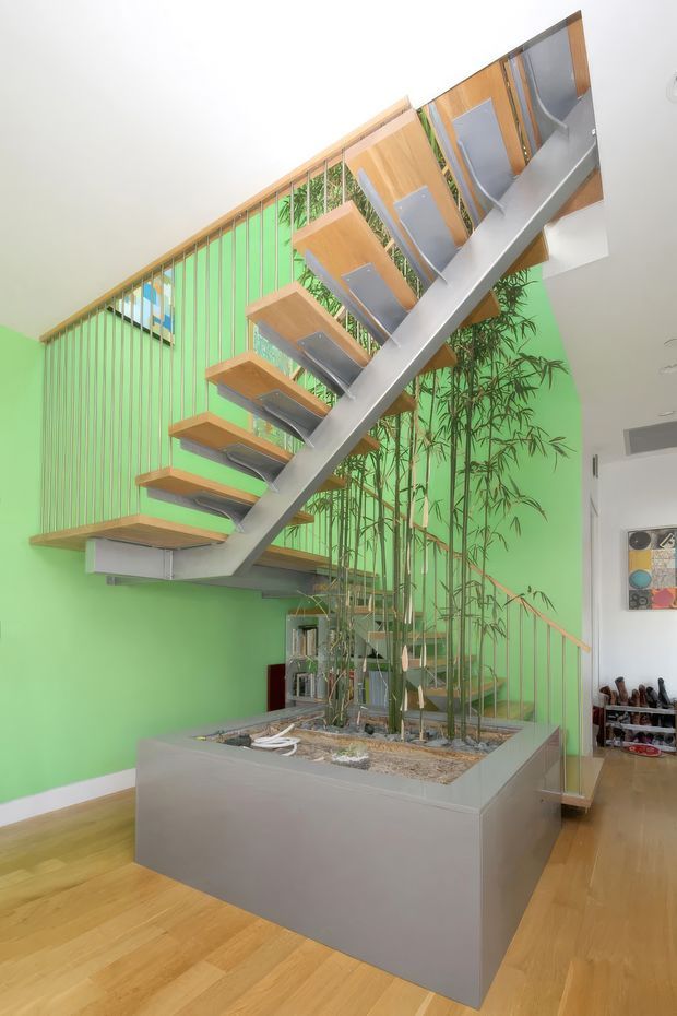 Interior view of a bright, contemporary stairway with floating timber treads supported by a prominent sloping metal spine. A large built-in planter filled with bamboo-like greenery sits beneath the stairs, backed by a vivid green wall with vertical timber slats. Timber flooring, glass balustrade elements, and an open upper-level landing create a dramatic sculptural focal point.