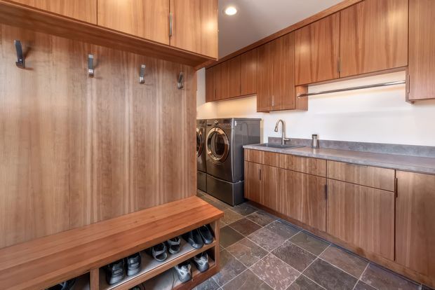 A compact, well-organised laundry and mudroom with extensive mid-tone timber cabinetry, a long stone-look benchtop, stainless sink and tap, integrated front-loading washer and dryer, and a built-in timber bench with open shoe storage. The room has dark patterned tile flooring, white walls, simple recessed lighting, and wall-mounted hooks.