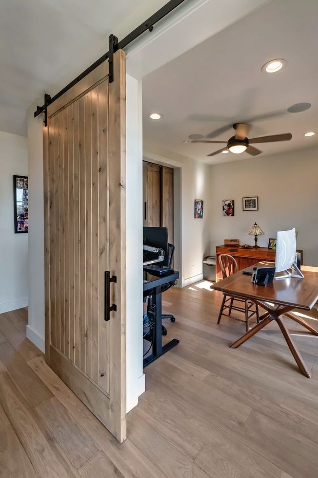 A bright home office viewed through a partially open barn-style sliding door. The room features light timber flooring, a large dark wood desk with crossed trestle legs, a wheeled office chair, a classic wooden sideboard, framed artwork, a table lamp, recessed ceiling lights, and a ceiling fan. The foreground door has vertical natural timber boards, black metal hardware, and a black overhead sliding track.