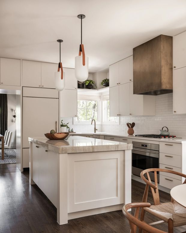 Bright, refined kitchen with a large central island, white shaker-style cabinetry, dark timber flooring, white subway-tile splashback, integrated stainless-steel appliances, and a warm stone benchtop. Two sculptural pendant lights hang above the island, while a small dining area with timber chairs is visible in the foreground. The kitchen has a clean, traditional-meets-contemporary character with natural timber accents.
