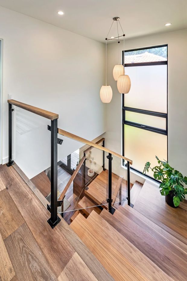 Bright contemporary stair landing viewed from above, featuring warm timber flooring and stair treads, a black metal and clear glass balustrade with timber handrails, a tall frosted-glass window, recessed ceiling lights, and three sculptural pendant lights. A potted plant adds greenery near the window.