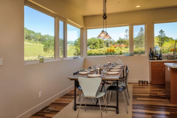 Bright, warm dining area adjoining a kitchen, with a long set dining table and mixed chairs positioned beside large corner windows. The room features timber flooring, light neutral walls, a suspended metal pendant light, and views onto a leafy landscaped setting.