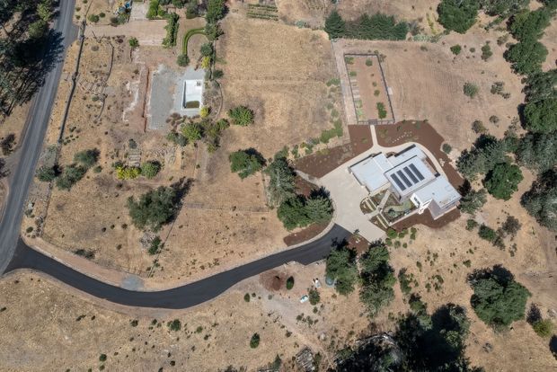 Aerial view of a large residential property in a dry, sparsely vegetated landscape. The light-coloured house has a broad pale roof with multiple skylights or roof windows, surrounding paved paths and garden beds, and a newly paved curved driveway connecting to a rural road. Scattered mature trees and landscaped areas sit within an expansive brown lot.