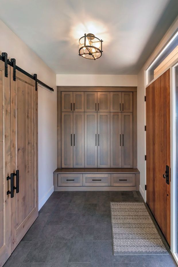 A narrow, well-organised entryway with extensive built-in timber cabinetry and a bench with drawers at the far end. A rustic sliding barn door with black metal hardware is visible on the left, while a timber entry door with surrounding glazing is on the right. The room has dark grey tiled flooring, warm neutral walls, and a decorative circular black-and-brass ceiling light.