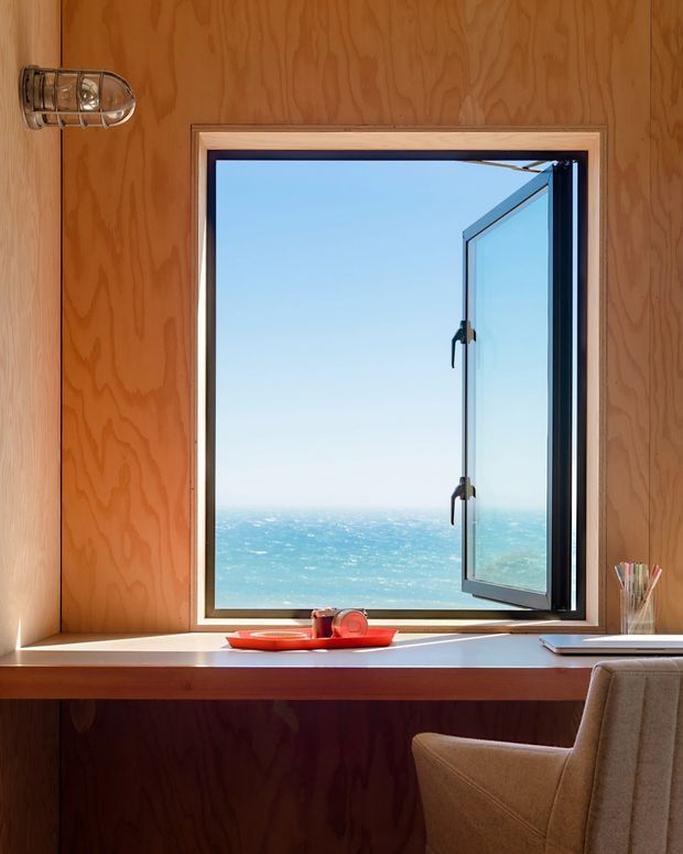 A compact wood-panelled interior workspace set against a large open casement window overlooking the ocean. Warm plywood wall and built-in desk surfaces frame the bright coastal view, with a simple wall light, stationery holder, red tray, and upholstered chair adding functional details.