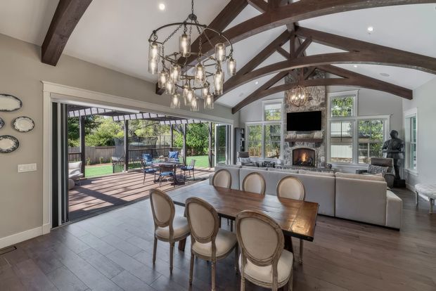 Open-plan dining and living area with a large timber dining table, upholstered dining chairs, and a sofa facing a fireplace and wall-mounted television. The room features dark exposed timber ceiling beams and trusses, a decorative multi-light chandelier, grey walls, and timber-look flooring. Large glazed sliding doors open onto a covered outdoor deck and landscaped garden.