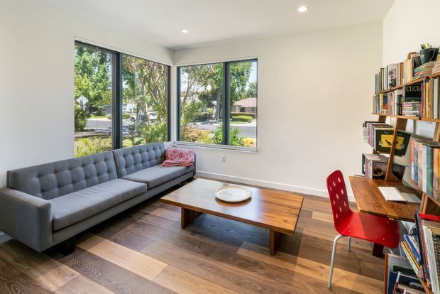Bright, compact study or living space with a grey tufted sofa, large corner windows, a wooden coffee table, and a wall-length open bookshelf incorporating a small desk. The room has dark timber flooring, white walls and ceiling, recessed downlights, and a red desk chair. The furnishings create a clean, contemporary and inviting atmosphere.