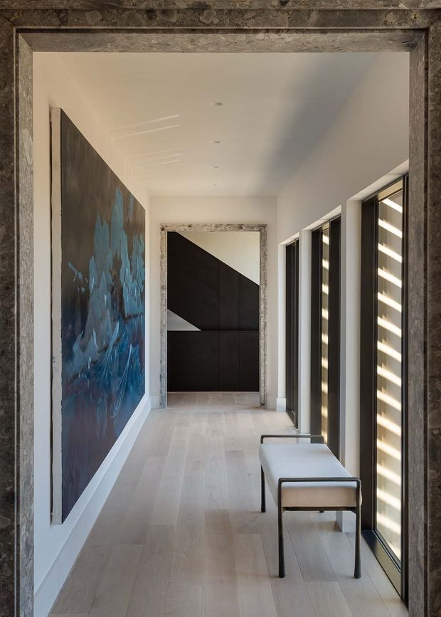 A refined, minimalist interior hallway viewed through a substantial dark stone or marble-framed opening. The corridor has pale timber flooring, white walls and ceiling, full-height dark-framed glazing along one side, and a simple upholstered bench. A large blue-and-charcoal abstract artwork lines the left wall, while a dramatic black geometric door or built-in feature anchors the far end. The space is bright, linear, and high-contrast with restrained contemporary detailing.