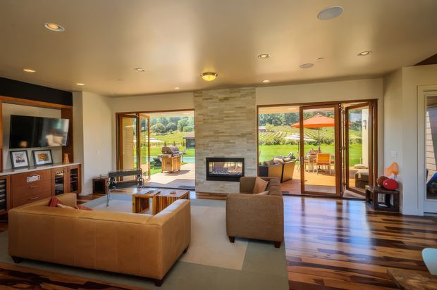 Spacious contemporary living room with a large tan sectional sofa, armchairs, built-in entertainment cabinetry, and a central stone-clad fireplace. Full-height glazed doors open to an outdoor entertaining area and landscaped grounds, while warm timber flooring contrasts with a pale carpeted seating zone. Recessed ceiling lighting and a simple pendant create a warm, inviting atmosphere.