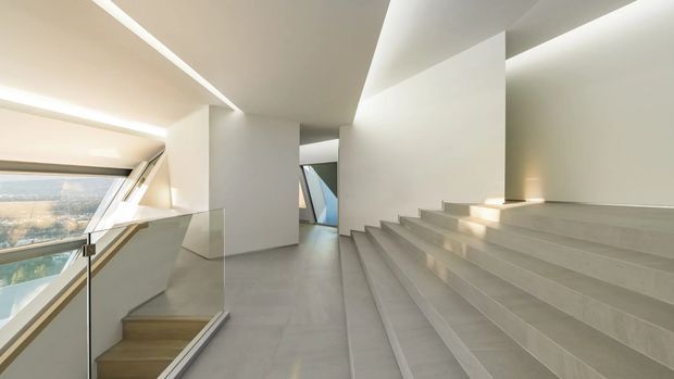 A bright minimalist interior stair hall with broad pale stone or concrete steps, a smooth grey floor, white sculptural walls, concealed linear lighting, and a glass balustrade. Large angled glazing on the left overlooks a distant urban landscape, while the staircase rises beside a generous open landing.