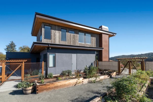 Exterior view of a contemporary two-storey house with a strong pitched roof, timber-lined upper facade, dark cladding, concrete-toned lower walls, and warm wood detailing. The property includes a landscaped garden, raised timber planter beds, fencing, and a pergola-like outdoor structure, with hills visible in the background.