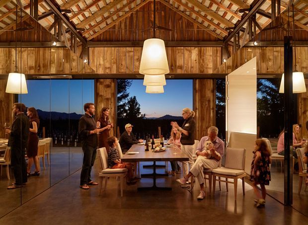 A warm, expansive dining and entertaining space with a long communal table, upholstered dining chairs, pendant lighting, and people gathered for a meal. The room features extensive timber wall cladding and a dramatic exposed timber roof structure with visible rafters and trusses. Full-height glazing opens views toward a dark mountainous landscape, while polished concrete flooring and black-framed glazing create contrast. A large white sculptural partition or fireplace-like feature stands to the right.
