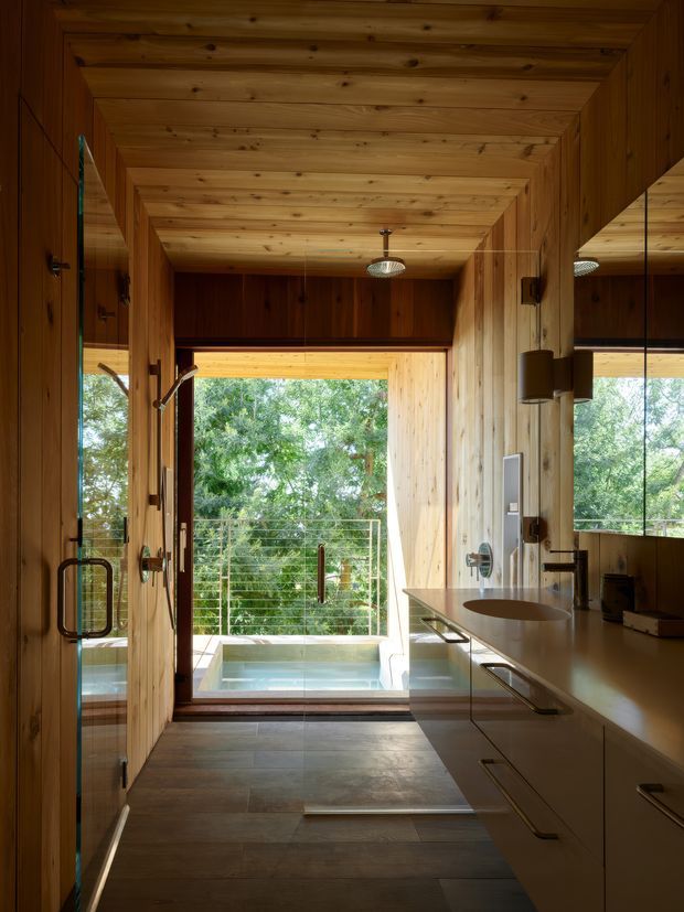 A warm, wood-lined bathroom with a long floating vanity and integrated basin on the right, a glass shower enclosure on the left, and a freestanding or partially visible bathtub near the glazed opening. The room opens to a small outdoor deck or balcony with a plunge pool and leafy greenery beyond. Timber cladding covers the walls and ceiling, while large mirrors and glazing amplify the compact space. The design is highly integrated and contemporary, with dark tiled flooring, streamlined cabinetry, pendant lighting, and natural daylight.