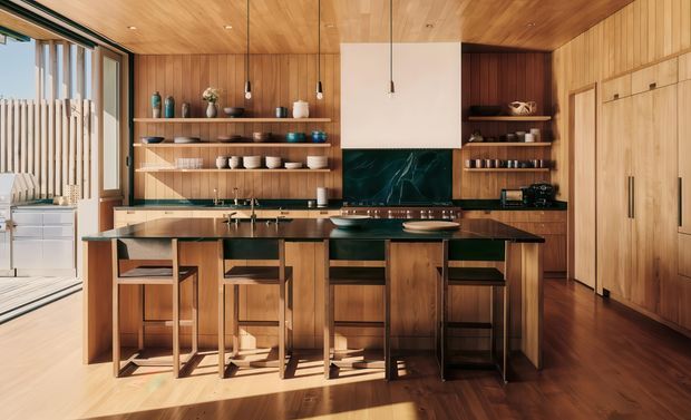 Warm, contemporary timber-lined kitchen with a large island and four bar stools, open shelving, integrated cabinetry, dark stone benchtops, and a deep green feature splashback. Timber ceiling, walls, cabinetry, and flooring create a cohesive natural interior, with generous glazing and an adjacent outdoor area visible at left.