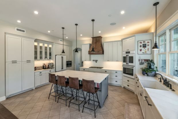 Bright, spacious traditional-style kitchen with extensive off-white cabinetry, a large central island with seating for four, integrated stainless-steel appliances, a tiled floor, white subway-tile backsplash, pendant lighting, and a farmhouse-style sink beneath large windows. The room has a warm, refined and inviting feel.