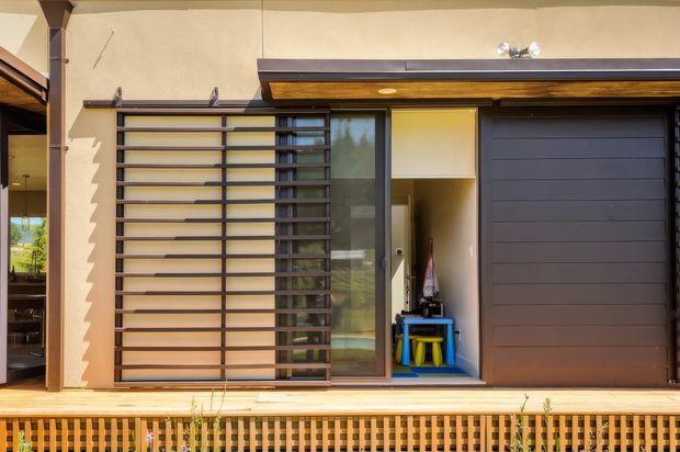 Exterior view of a contemporary residential facade featuring a dark sectional garage door, glazed entry or living-area door, horizontal metal screening, warm timber soffit, and a timber deck with a lattice balustrade. Neutral rendered walls and strong horizontal lines create a clean, modern appearance.