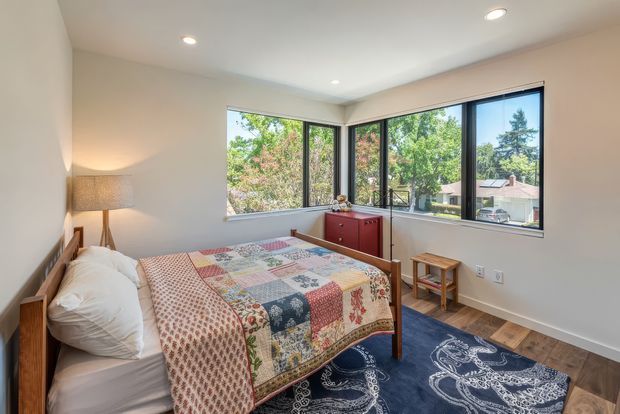 A bright, compact bedroom with a wooden bed, colourful patchwork quilt, navy patterned area rug, small wooden bedside tables, and a red timber dresser. Large corner windows provide abundant natural light and views of leafy suburban surroundings. The room has white walls, recessed ceiling lights, and dark timber flooring.
