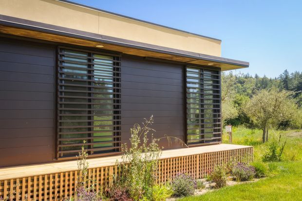 Single-storey modern building facade with dark horizontal cladding, broad roof overhang, and two large windows screened by dark horizontal louvres. A timber deck with patterned vertical balustrade runs along the front, bordered by landscaped planting and a rural, tree-filled setting.