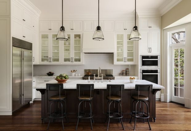 A refined, spacious kitchen with a large central island and seating for five. The room features white raised-panel cabinetry with glass-front display cupboards, dark wood island cabinetry, a pale stone benchtop, stainless-steel appliances, a tiled backsplash, pendant lighting, and timber flooring. Tall French doors and a high ceiling bring in natural light.